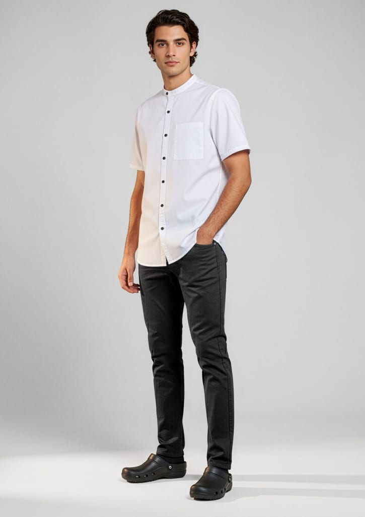 A man stands wearing black chef flex pants paired with a short-sleeve white shirt. The outfit includes a logo.