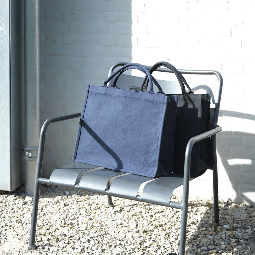 Navy blue recycled tote bags are displayed on a metal chair against a light brick wall. One bag is slightly behind the other.